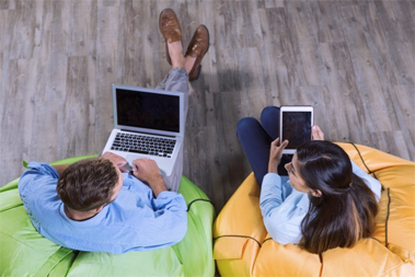 um homem usando laptop sentado em um puff verde conversando com uma mulher usando tablet sentada no puff amarelo